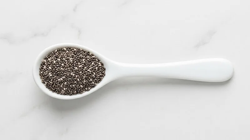 Two tablespoons of chia seeds measured in a white ceramic spoon on marble counter